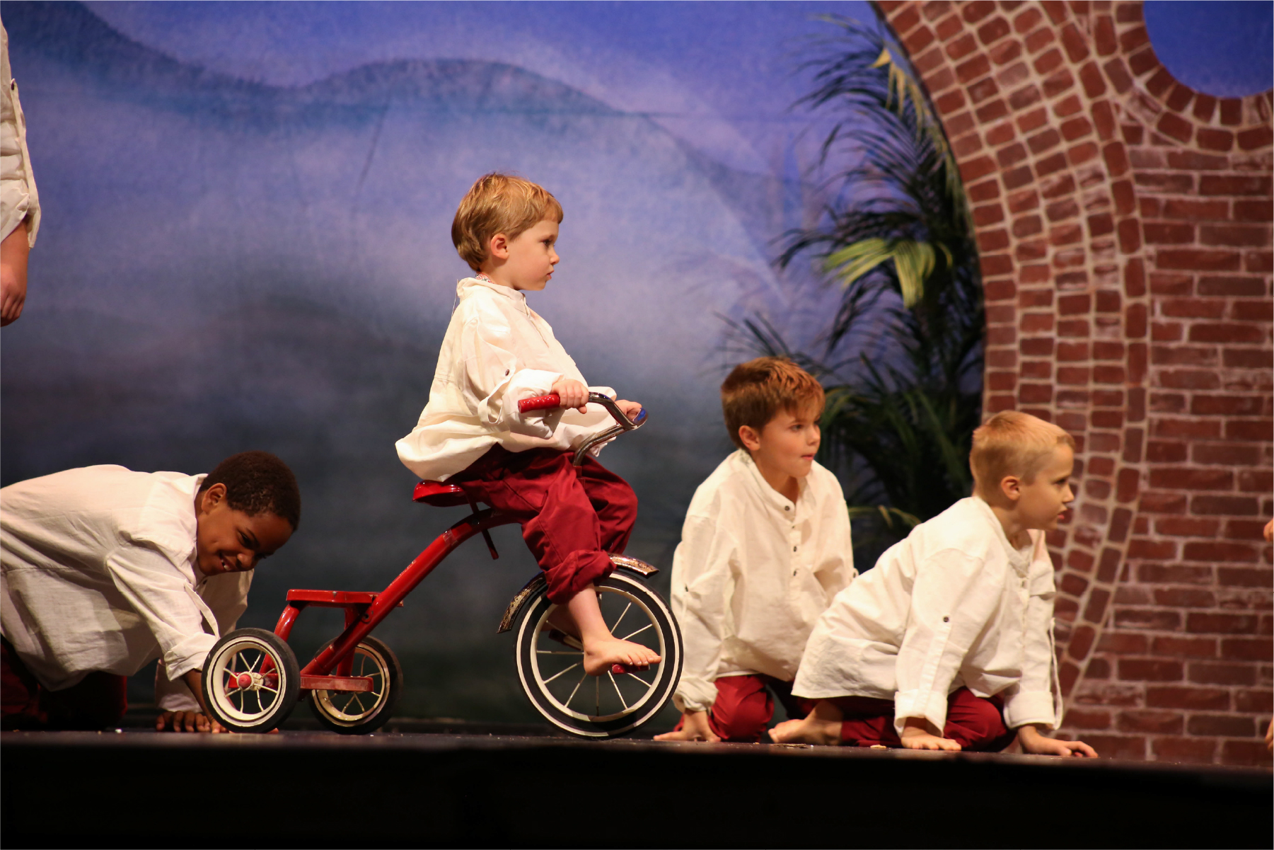 Four young boys acting as characters in the story of Amy Carmichael. One boy I son a tricycle while the other boys are looking off stage
