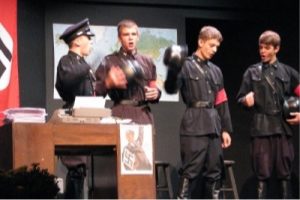 Group of young actors dressed in period clothing as Nazi guards on stage.