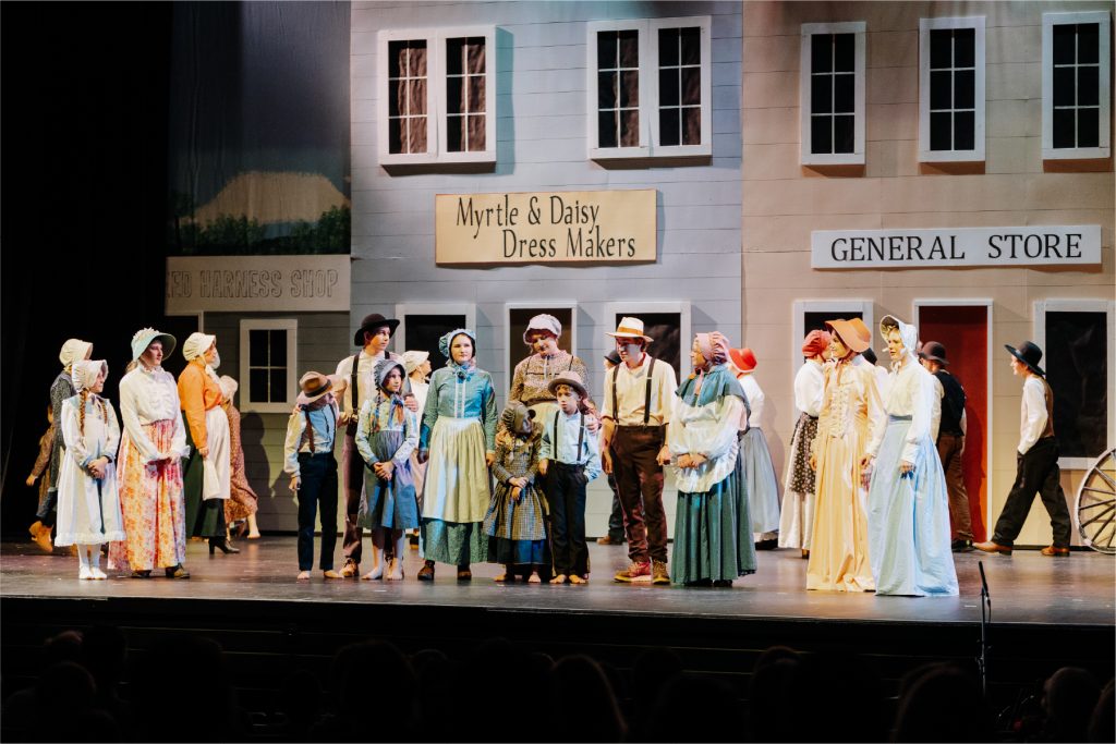 Young actors and actresses milling about on stage dressed in period costume for a plat set on the Oregon Trail