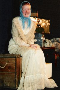 Young actress dressed in a white period dress with a scarf around her head sitting on a box smiling at the camera.