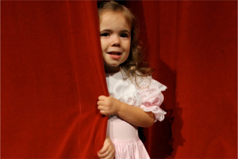 Young girl in a pink dress peeking out of the red stage curtain smiling