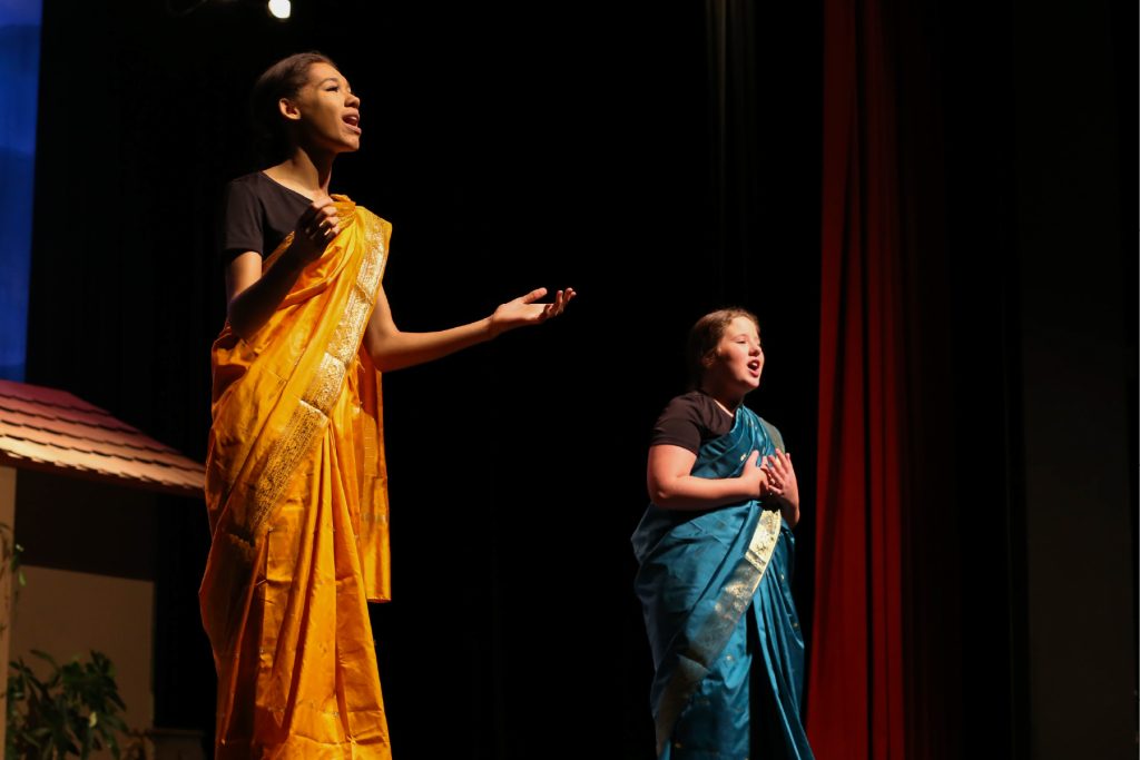 Two young actresses on stage in period costumes from India singing.